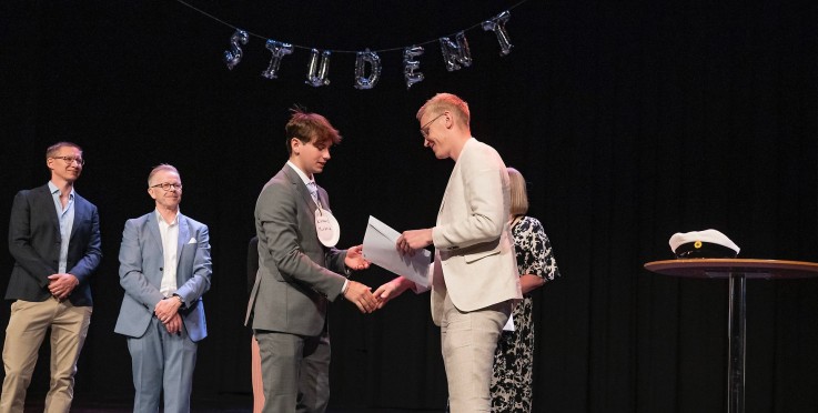 Personer i kostym på scen, en student får diplom, ballonger bildar ordet 'STUDENT', studentmössa på bord.