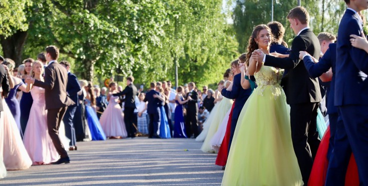 Ungdomar i festkläder dansar parvis utomhus på en solig dag, omgivna av gröna träd och åskådare.