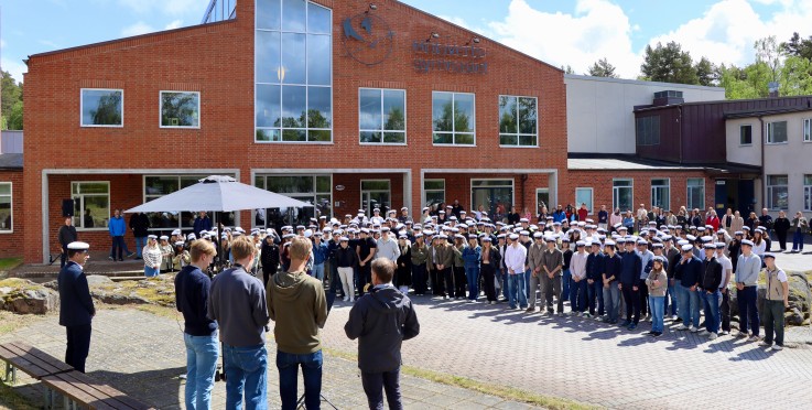 Stor grupp människor samlade utanför en röd tegelbyggnad, många bär vita studentmössor.