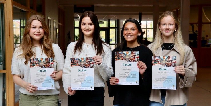 Fyra personer står inomhus och håller diplom, deras ansikten är suddade.