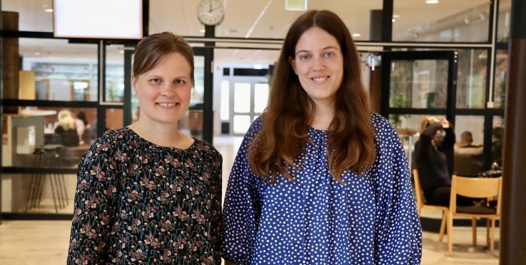 Två personer står inomhus i en modern miljö, ansikten är pixlade, bakgrund med glasväggar och klocka.