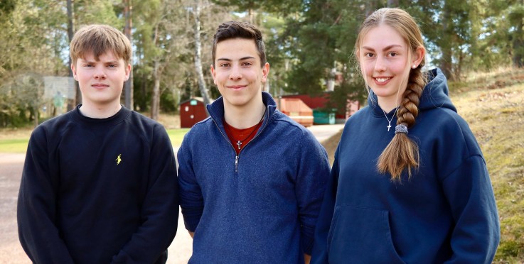 Tre personer står utomhus i en park, ansiktena är suddade. De bär mörka tröjor och korshalsband.