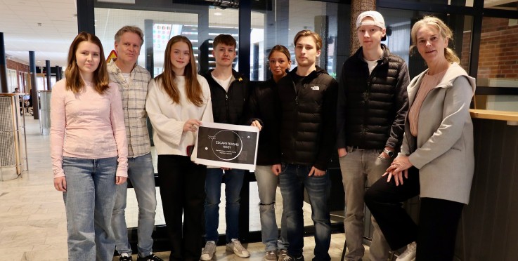 En grupp människor står tillsammans inomhus, en person håller en skylt med texten 'Escape Rooms Hogy'.