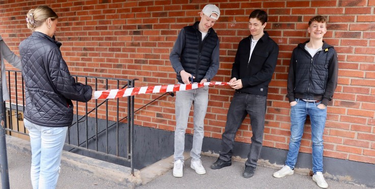 Fyra personer står vid en röd tegelvägg och håller ett rödvitt avspärrningsband.