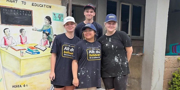 Fyra personer står framför en vägg med en målning om utbildning, klädda i svarta t-shirts och kepsar.