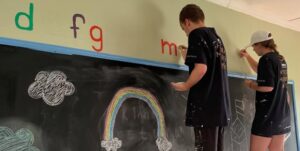 Två personer målar bokstäver på en vägg ovanför en svart tavla med kritteckningar av regnbåge och moln.