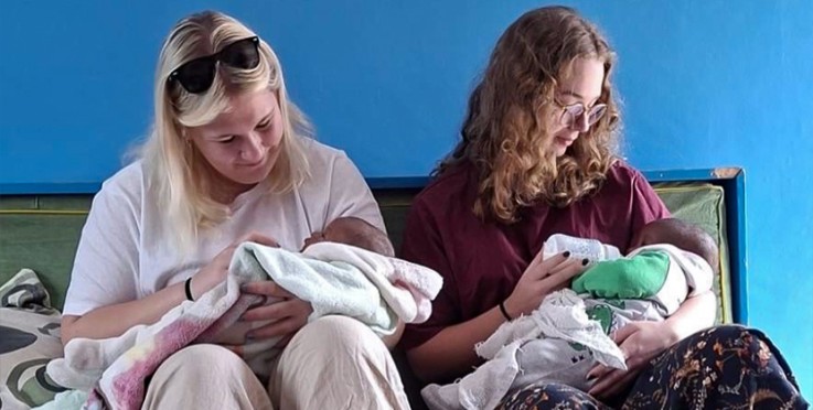 Två kvinnor sitter på en soffa och håller varsin baby i famnen, mot en blå vägg.