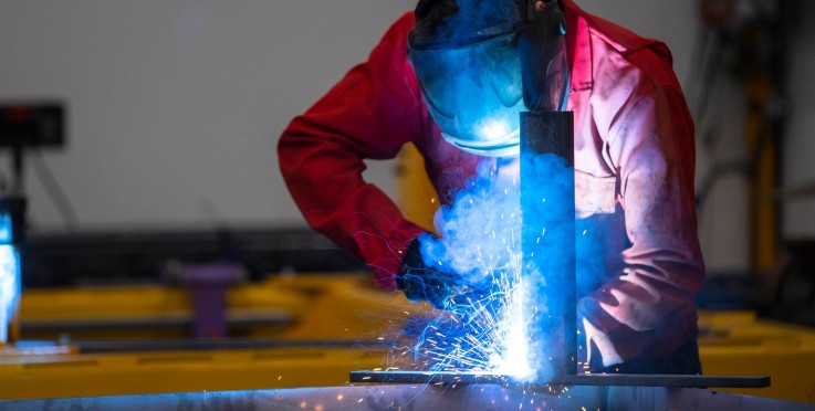 En person i röd skyddsdräkt och visir svetsar metall med starka gnistor och blå rök.