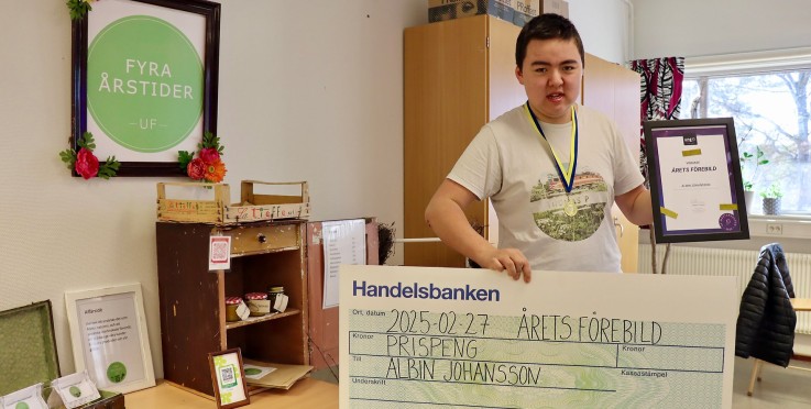 Person håller en stor check och ett diplom i ett klassrum, bredvid ett UF-företags utställning.