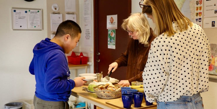 Tre personer står vid ett bord och serverar fika med bullar, koppar och tallrikar i ett rum med anslag på väggarna.