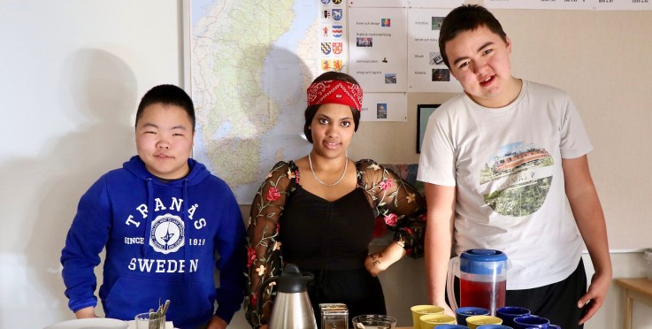 Tre personer står vid ett bord med muggar och en kanna, framför en karta och affischer på väggen.