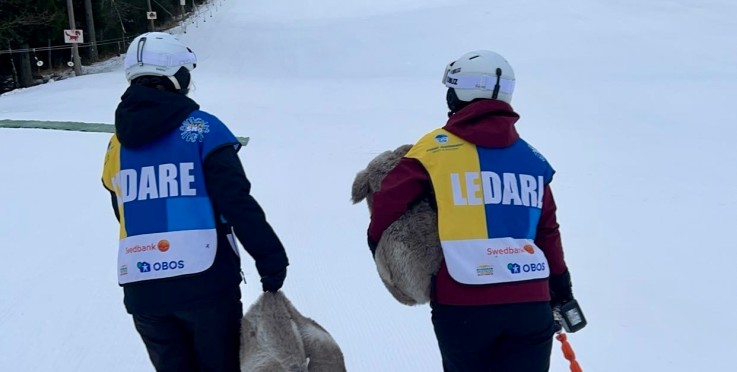 Två personer med hjälm och väst med texten 'LEDARE' går på en snötäckt skidbacke, en bär ett gosedjur.