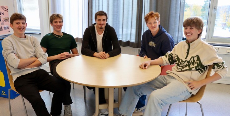 Fem unga personer sitter runt ett runt bord i ett ljust rum med stora fönster och gardiner.