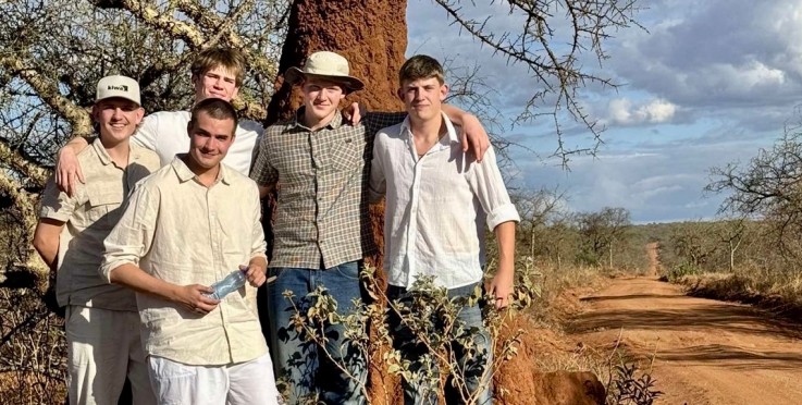 Fem personer står tillsammans vid ett stort termitbo på en grusväg i savannliknande miljö.