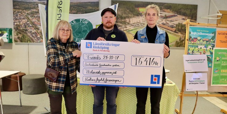 Tre personer håller en stor check på ett event, omgiven av informationsskyltar och affischer.