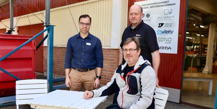 Tre personer, två stående och en sittande, utanför en byggnad med skyltar och ett vitt bord.