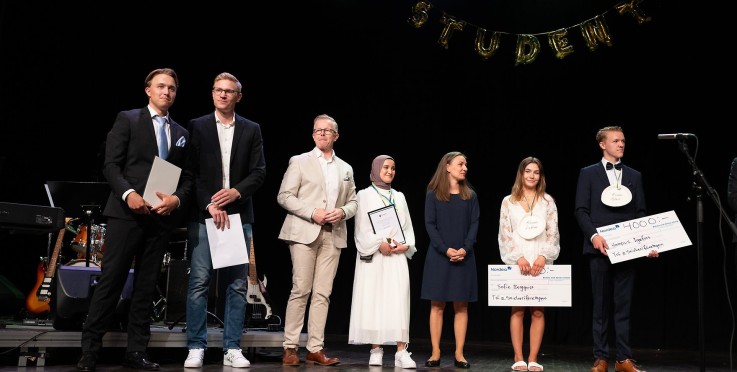 Flera personer står på en scen under en studentceremoni, några håller diplom och stora checkar.
