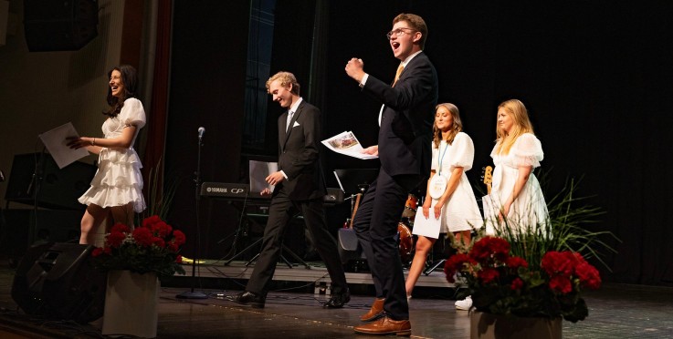 Flera personer står på en scen, klädda i festkläder, med papper i händerna. Blommor och instrument syns.