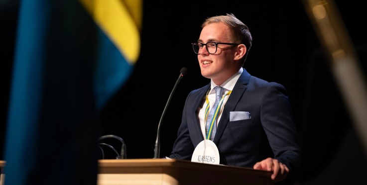En person i kostym står vid ett podium och talar, med en medalj runt halsen och en svensk flagga i förgrunden.