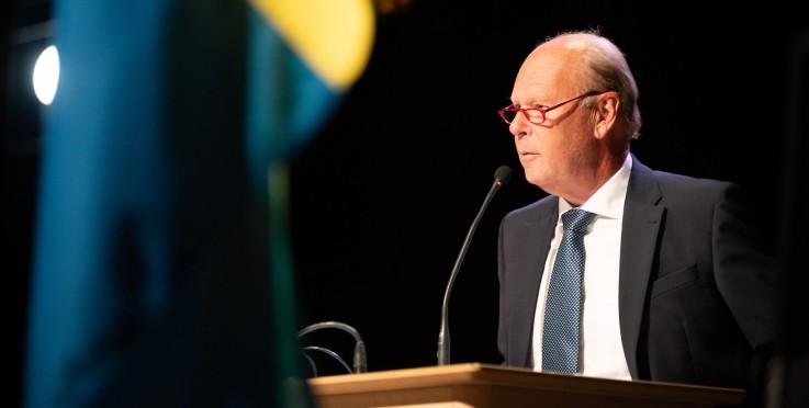 Man i kostym talar vid podie med mikrofon, svensk flagga syns i förgrunden.