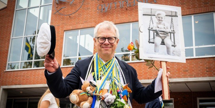 Student firar examen utanför skola med blommor, gosedjur och skylt med barndomsfoto.