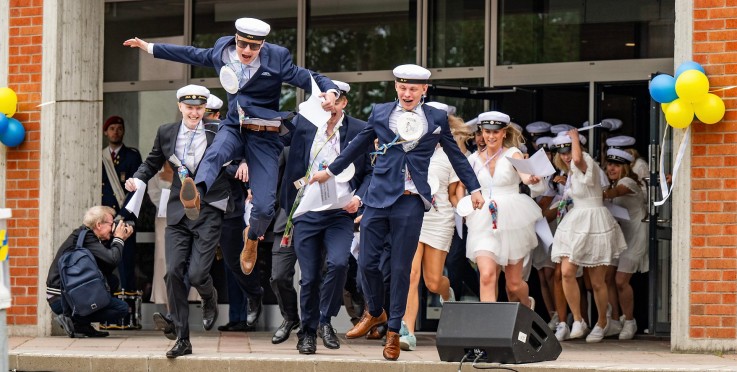 Studenter i vita mössor springer ut från en skolbyggnad, firar examen med glädje och ballonger i bakgrunden.