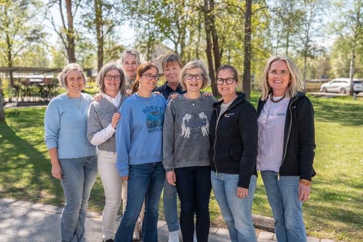 En grupp på sju personer står tillsammans utomhus i en park med träd och gräs i bakgrunden.