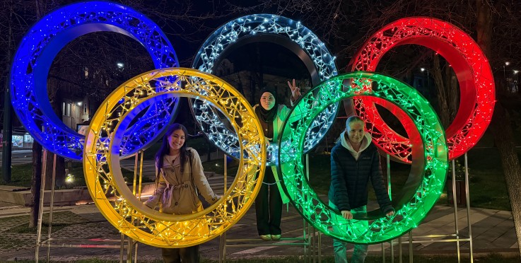 Tre personer poserar framför upplysta olympiska ringar utomhus på natten.