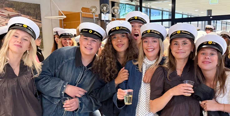 En grupp unga personer i studentmössor står tillsammans inomhus och firar, flera håller i glas.