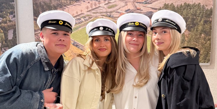 Fyra personer med studentmössor poserar tillsammans inomhus framför en tavla med flygbild.