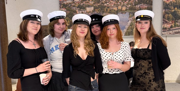 En grupp unga personer med studentmössor poserar inomhus framför en vägg med flygbilder.