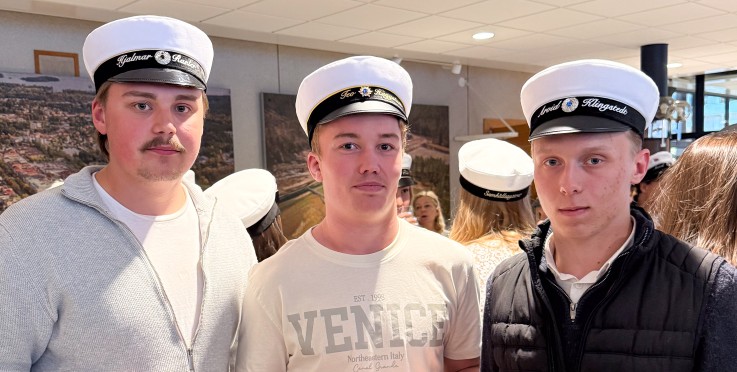 Tre unga män med studentmössor står tillsammans inomhus och håller drycker i händerna.