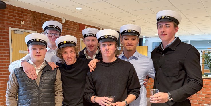 Åtta unga män med studentmössor står tillsammans inomhus och poserar för ett gruppfoto.