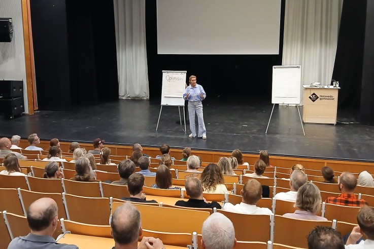 Föreläsare står på scen framför publik i en aula med stor projektorduk och blädderblock.