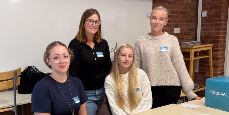 Fyra personer sitter och står vid ett bord i ett klassrum med papper och listor framför sig.