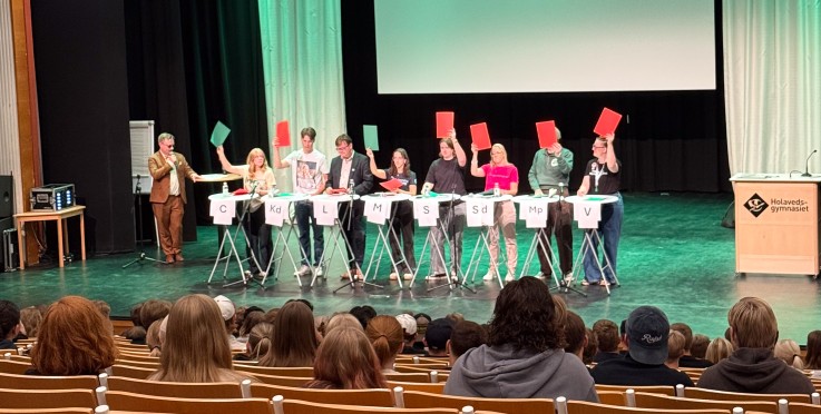 Debatt på scen med flera personer som håller upp röda och gröna skyltar inför publik i en aula.