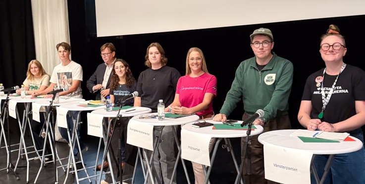 Åtta personer står bakom ståbord på en scen, redo för en paneldebatt. Namnskyltar visar olika partier.