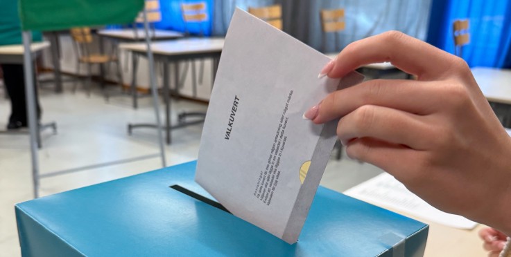En hand lägger en valsedel i en blå valurna märkt 'SKOLVAL' i ett klassrum.
