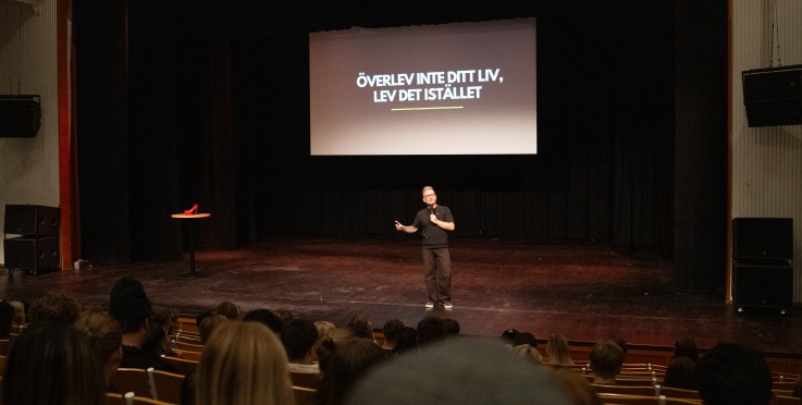 Föreläsare står på scen framför publik med texten 'Överlev inte ditt liv, lev det istället' projicerad bakom sig.