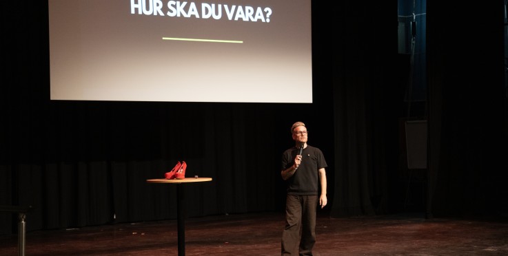 En man står på en scen med mikrofon, bredvid ett bord med röda högklackade skor. Text på skärmen: 'Hur ska du vara?'.