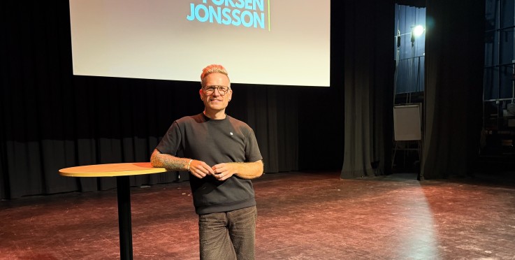 En person står på en scen bredvid ett runt bord, med en presentation på stor skärm bakom sig.