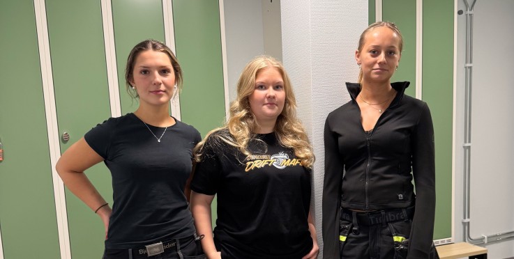 Tre unga kvinnor står i ett omklädningsrum med gröna skåp, klädda i arbetskläder.