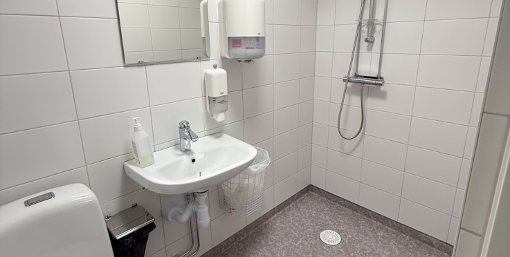 Ett vitt, handikappanpassat badrum med toalett, handfat, spegel och dusch utan duschkabin.