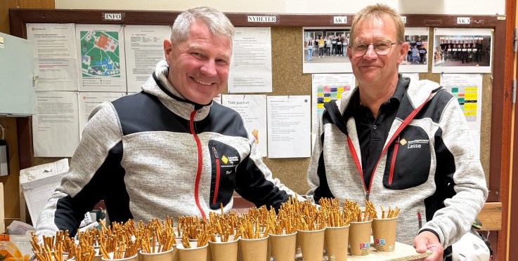 Två män i arbetskläder håller en bricka med pappersmuggar fyllda med salta pinnar i ett personalrum.