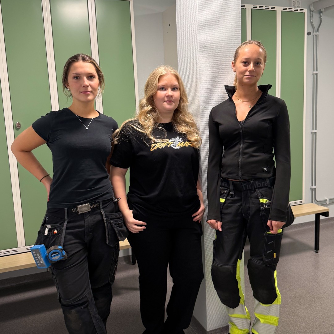 Tre unga kvinnor står i ett omklädningsrum med gröna skåp, klädda i arbetskläder.