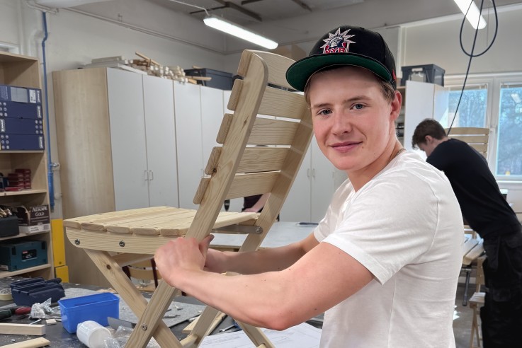 Ung man bygger en trästol i ett ljust snickeri, bär vit t-shirt och svart keps.