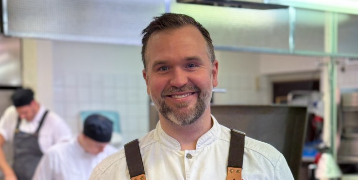 Leende kock i röd förkläde står i restaurangkök med andra kockar i bakgrunden.