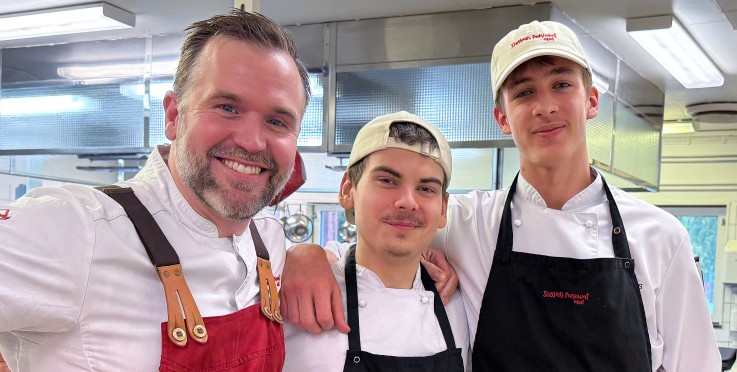 Tre kockar i arbetskläder poserar tillsammans i ett restaurangkök och ler mot kameran.