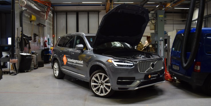 En grå Volvo SUV med öppen motorhuv står i en verkstad. Texten 'Holavedsgymnasiet' syns på sidan.