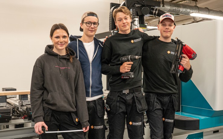 Fyra unga personer i arbetskläder står framför en industrimaskin, två håller verktyg och alla ler mot kameran.